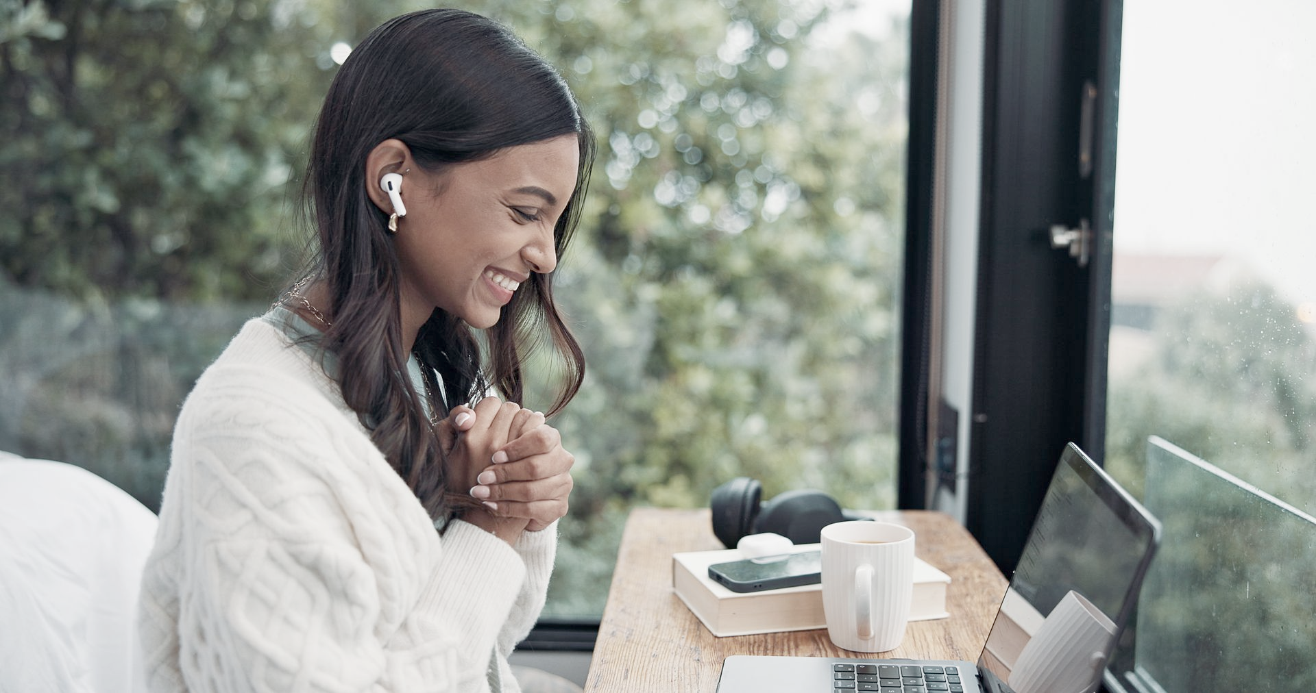 Woman smiling, wearing earbuds, working on laptop by a window, with coffee cup and headphones on desk.