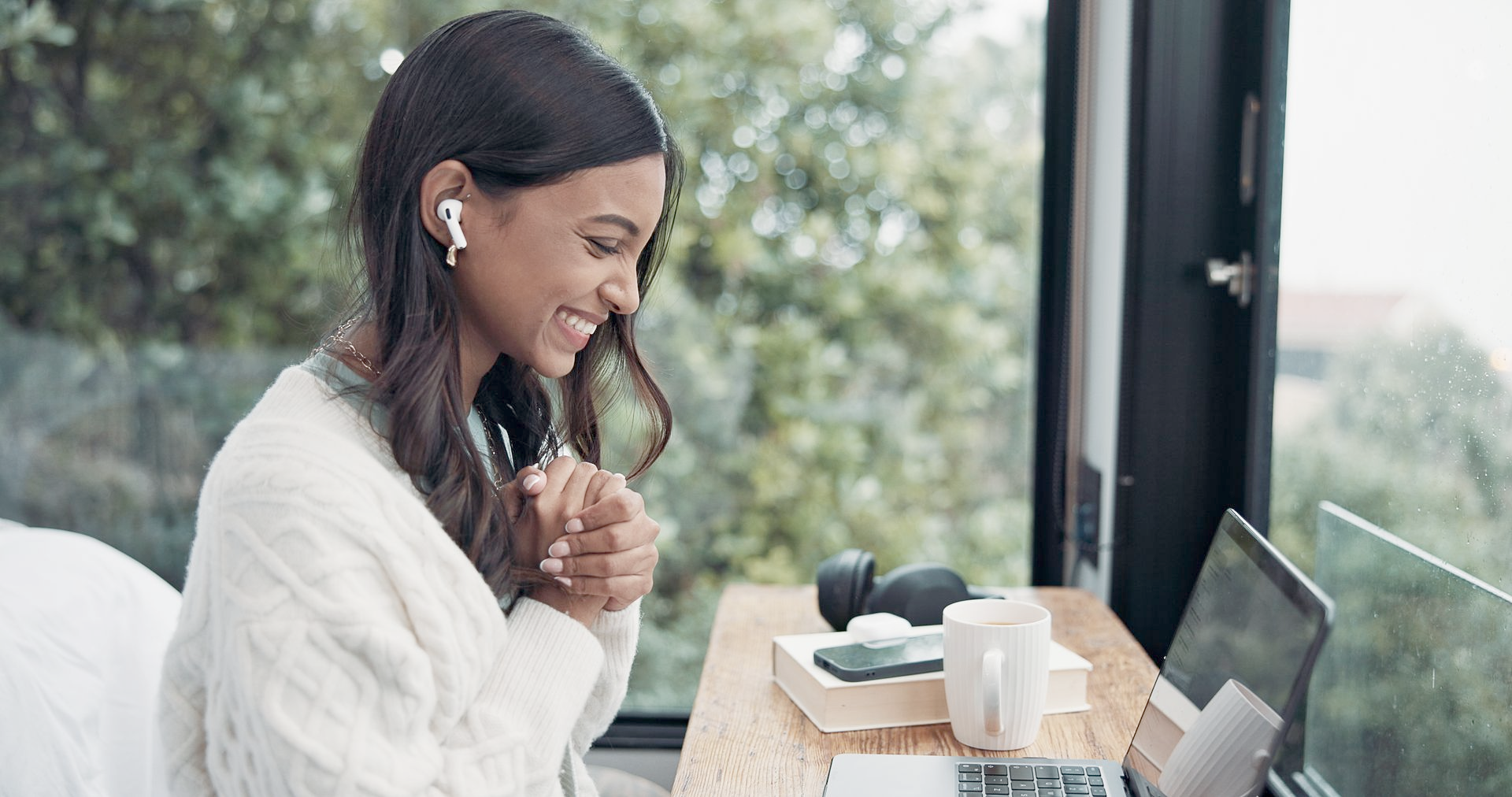 Woman in sweater smiles during video call, working at wooden desk near a window.