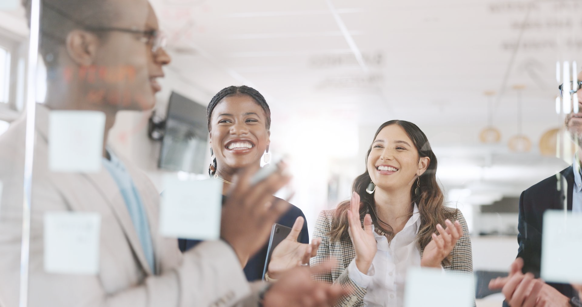 Group of people clapping, smiling, in a meeting room, near a glass wall with sticky notes.