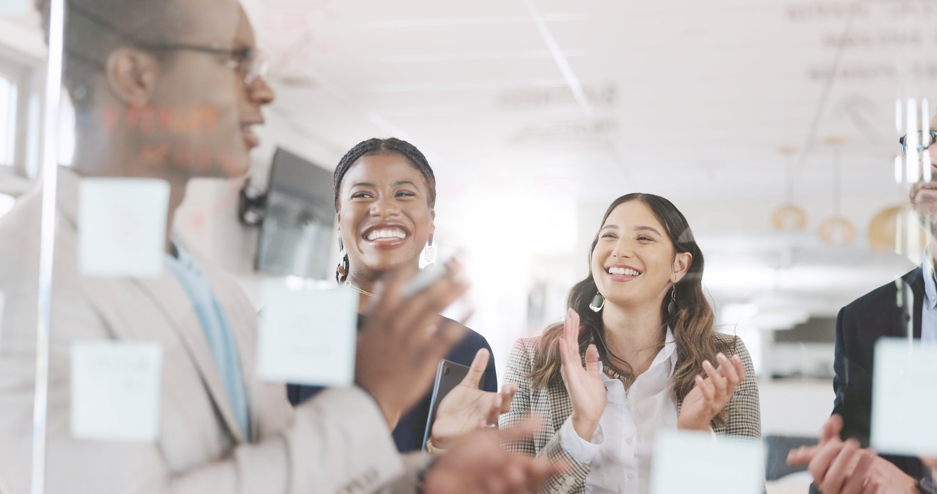 Group of people in office, applauding and smiling during a presentation, with sticky notes on the glass.