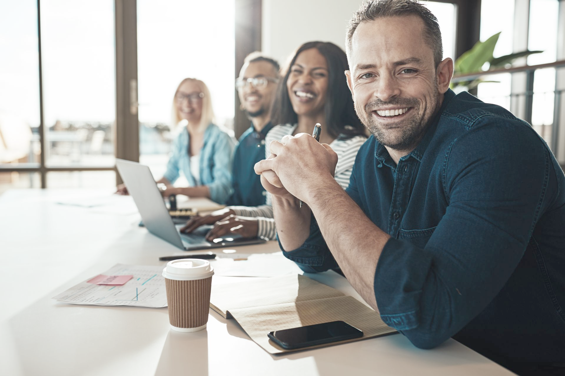 Group of smiling professionals at a table, working on laptops, with a coffee cup.