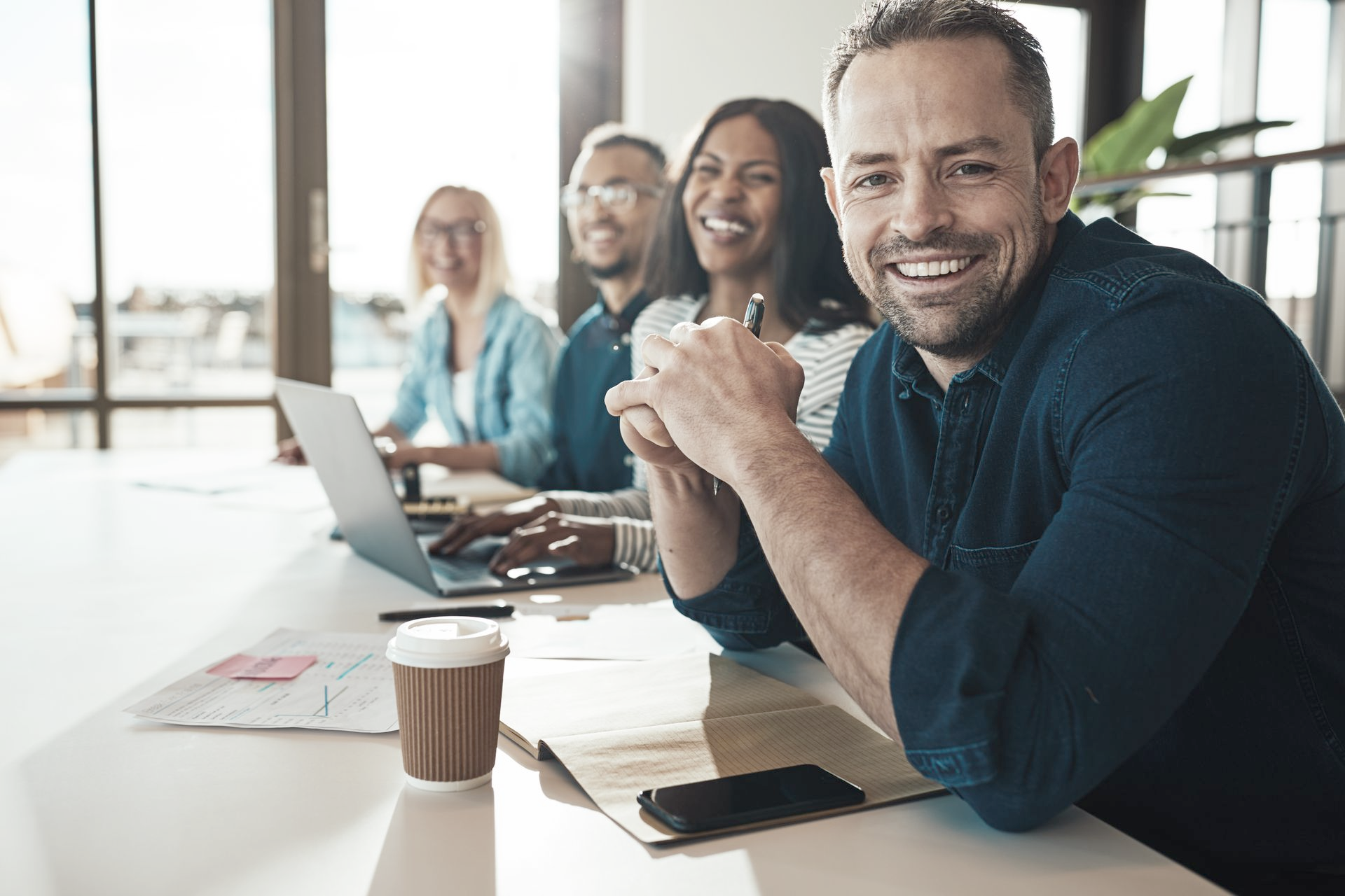Group of diverse colleagues laughing around a table in an office, with laptops and coffee.