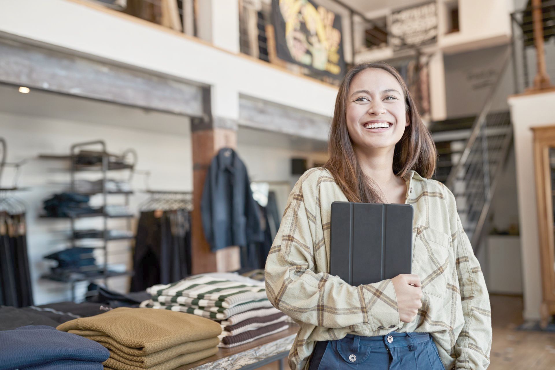 Woman in clothing store, smiling, holding tablet, standing near a display of folded sweaters.