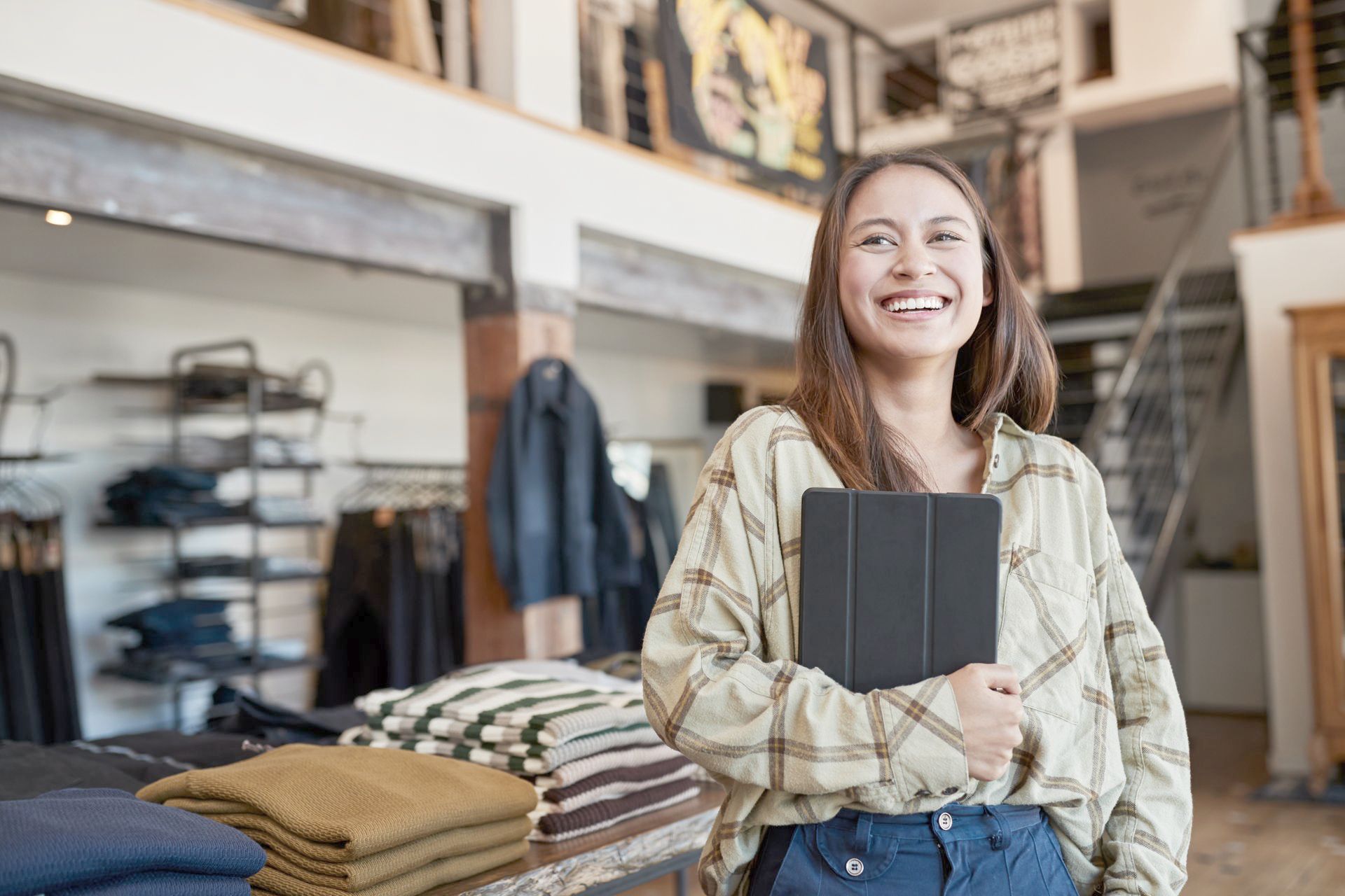 A woman is holding a tablet in her clothing boutique.