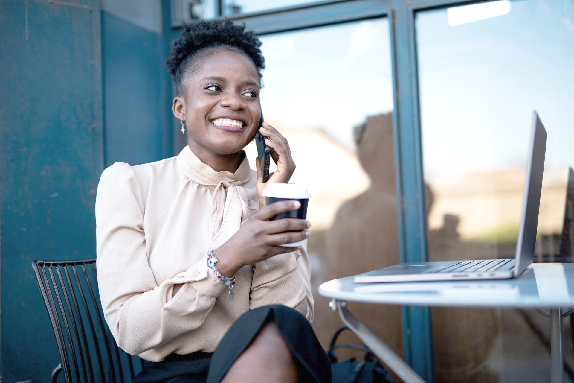Woman on phone, smiling, at table with coffee and laptop, outdoors with blue wall.