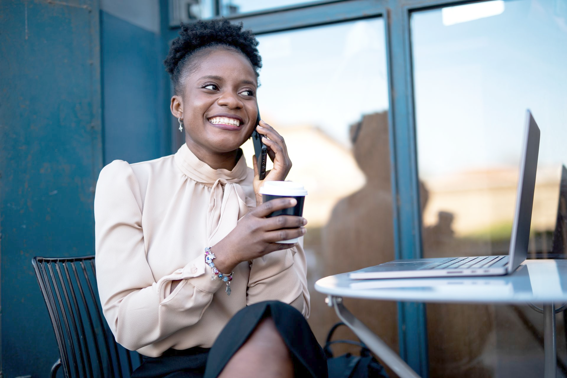Woman smiling, talking on phone, holding coffee, using laptop at outdoor table.