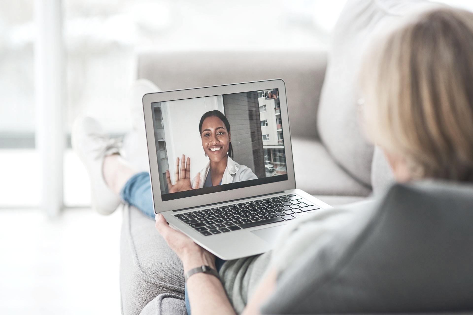 Woman reclines on a sofa, laptop in hand, video chatting with a smiling Black doctor indoors.