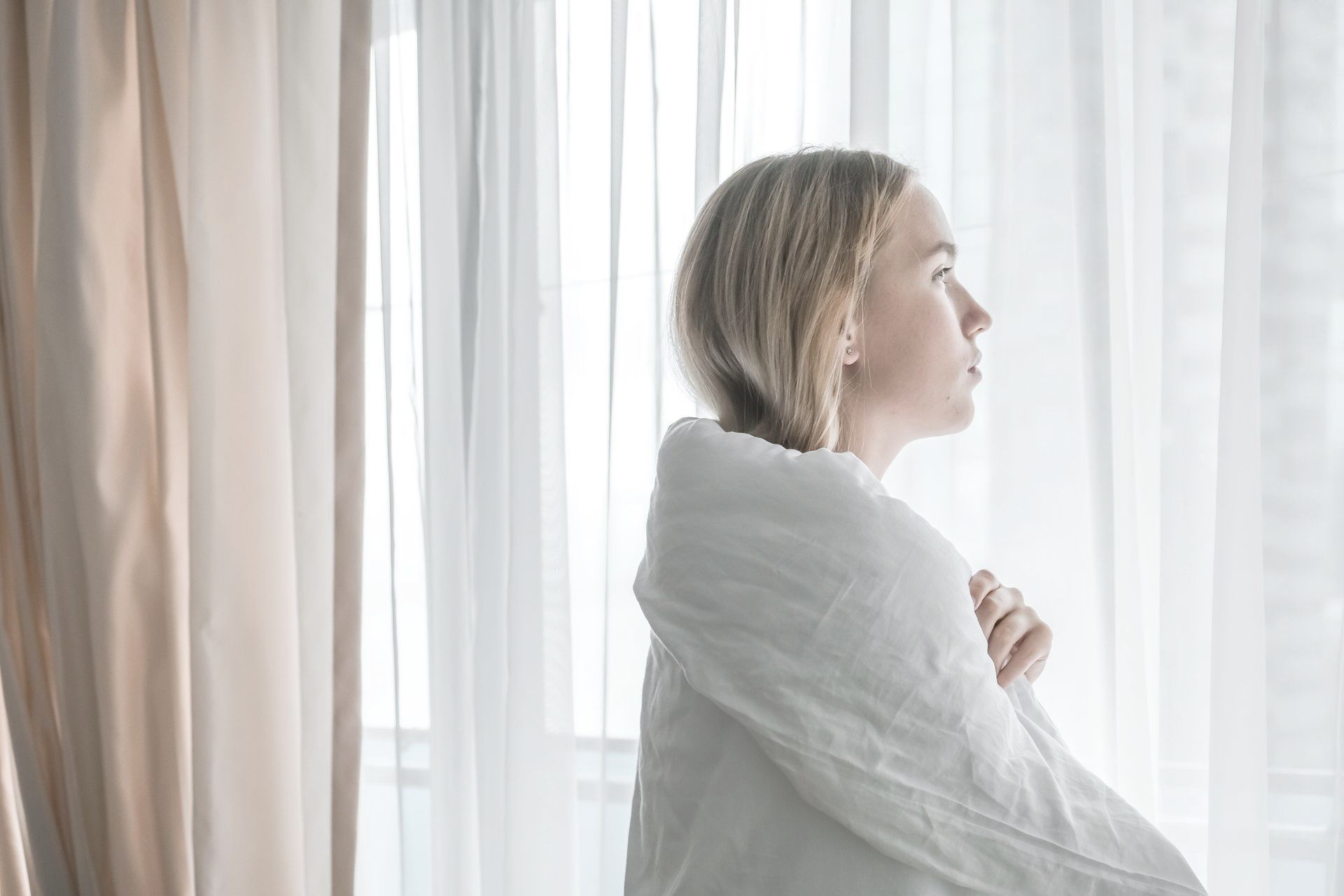 A woman wrapped in a white blanket is looking out of a window.