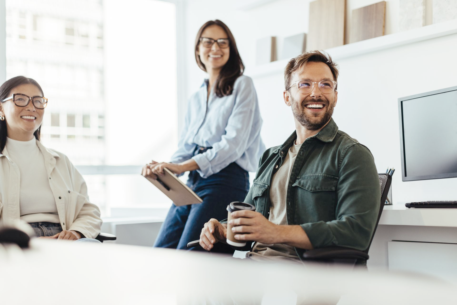 Three smiling coworkers in a bright office: one holding a coffee, one with a tablet, one at a desk.