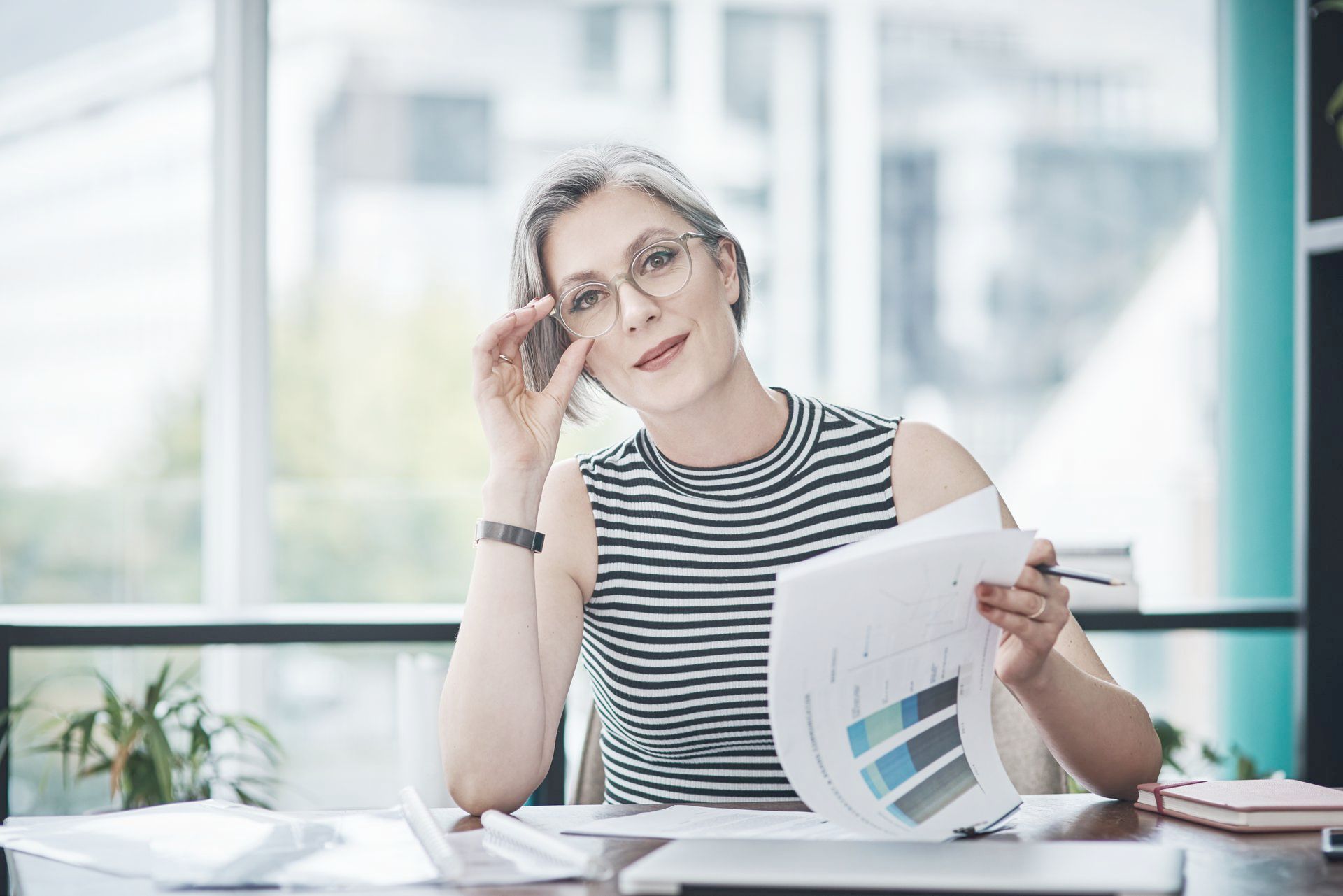 A woman is sitting at a desk holding a piece of paper and talking on a cell phone.