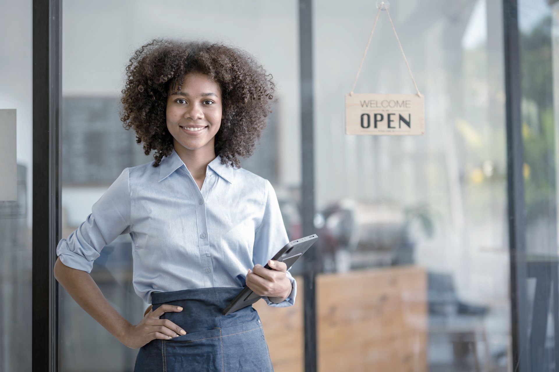 A woman is standing in front of a glass door with an open sign holding a tablet.