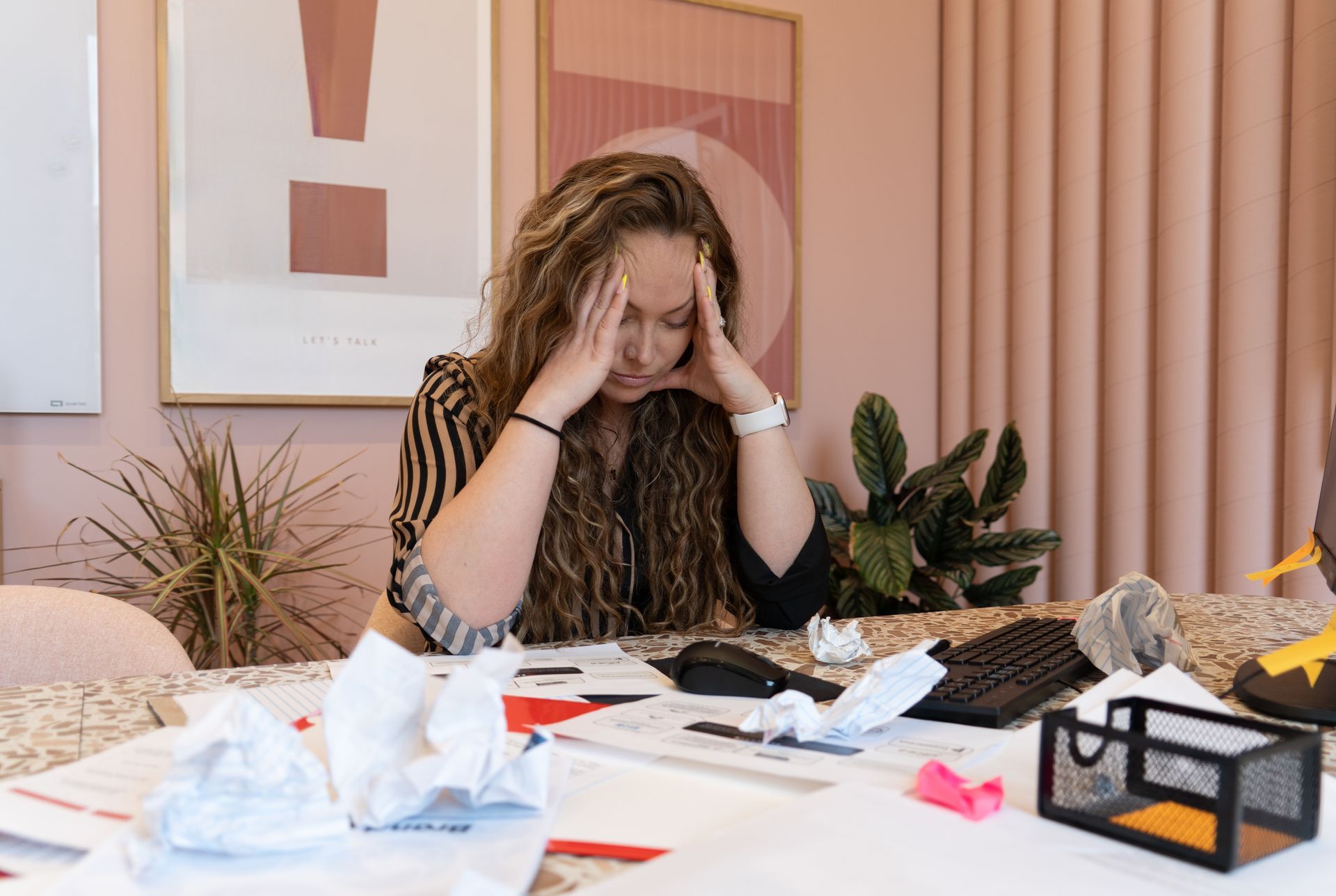 Woman with hands on head surrounded by papers, appearing stressed, in her office.