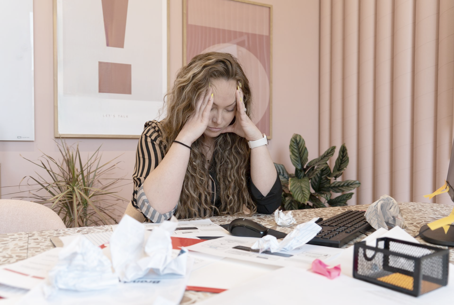 Woman with head in hands, stressed at messy desk with crumpled paper and calculator.
