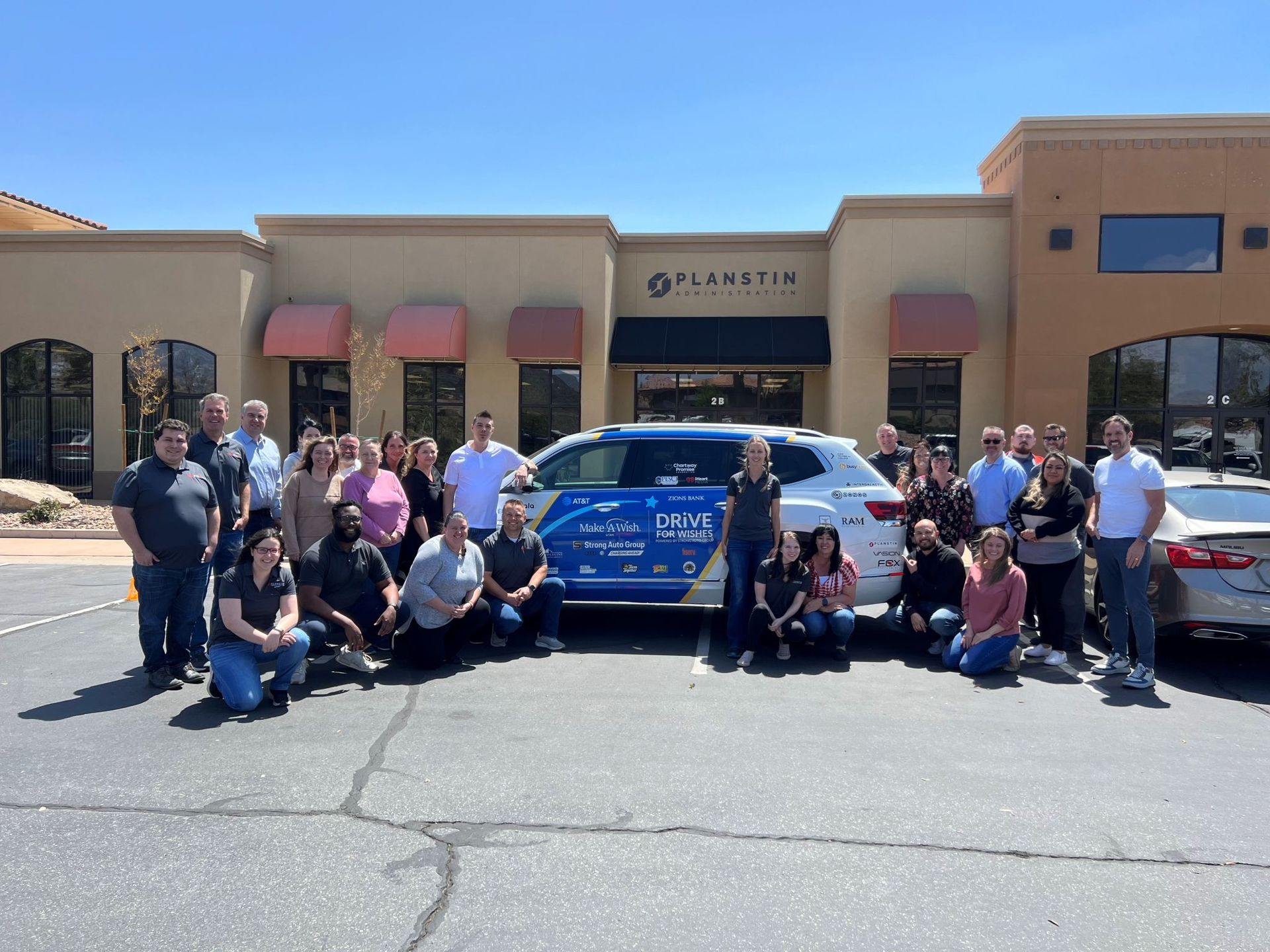 A group of people posing for a picture in front of a building with the Make-A-Wish car.