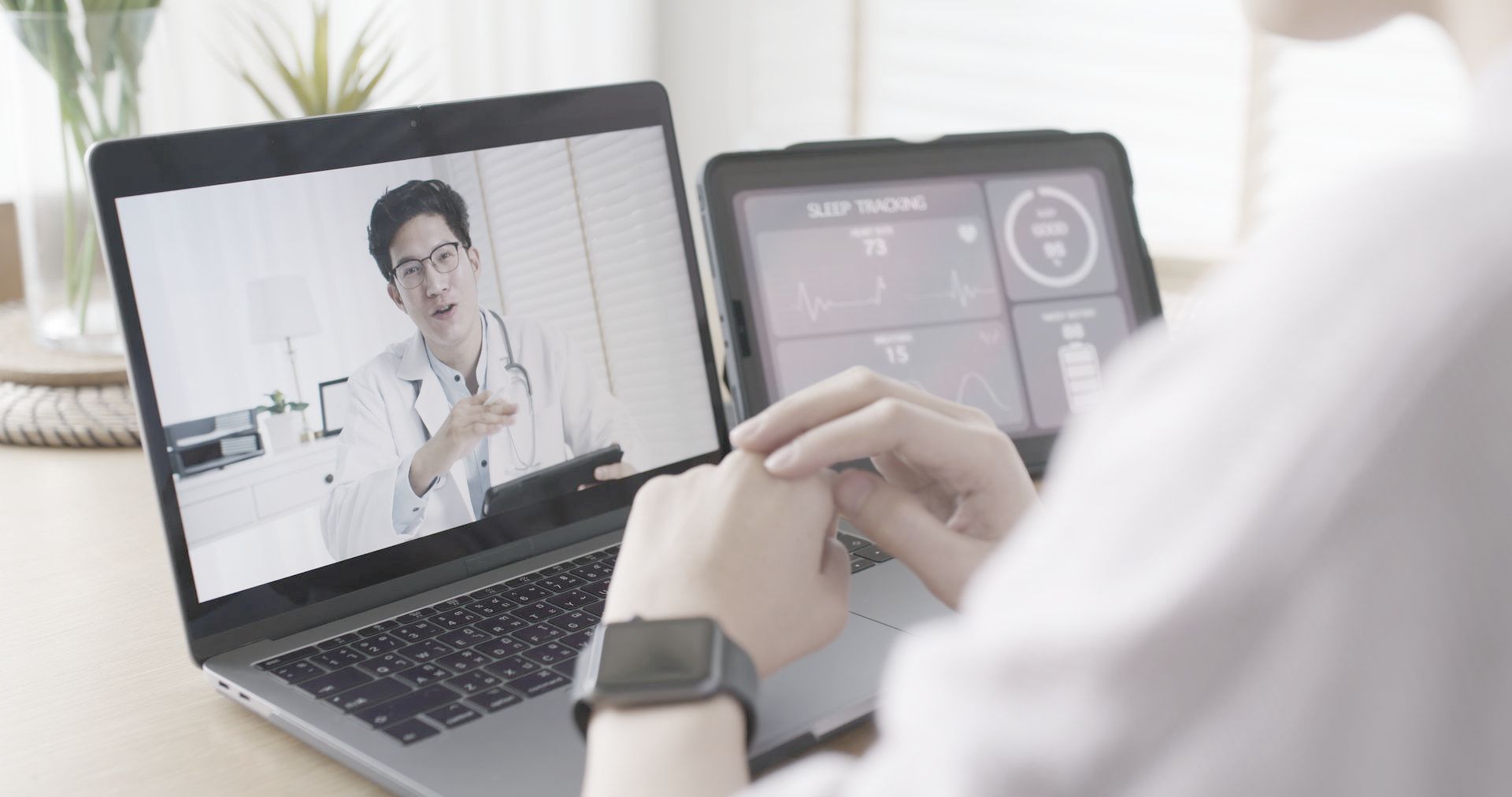 A person is using a laptop and a tablet to have a video call with a doctor.