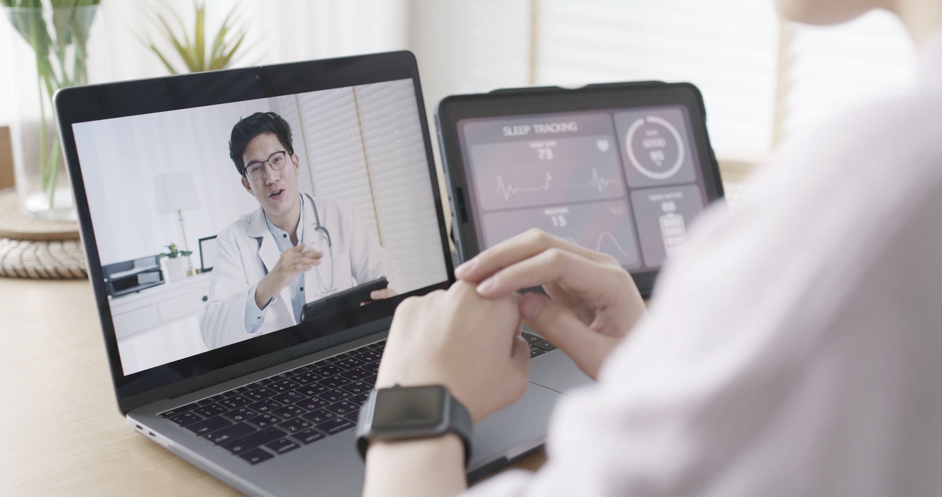 A person is sitting at a table using a laptop and a tablet to talk to a doctor.