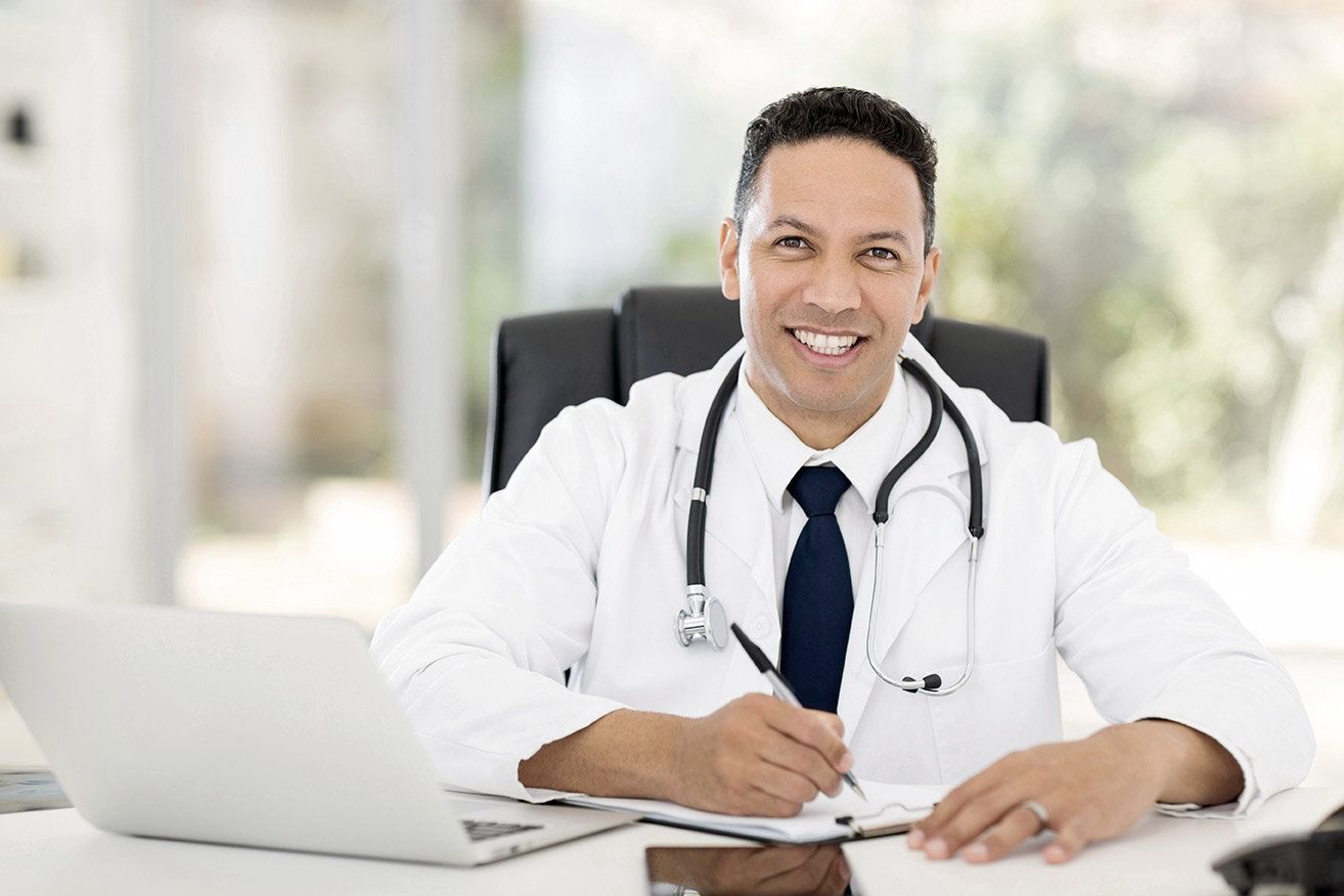 A doctor is sitting at a desk with a laptop and a clipboard.