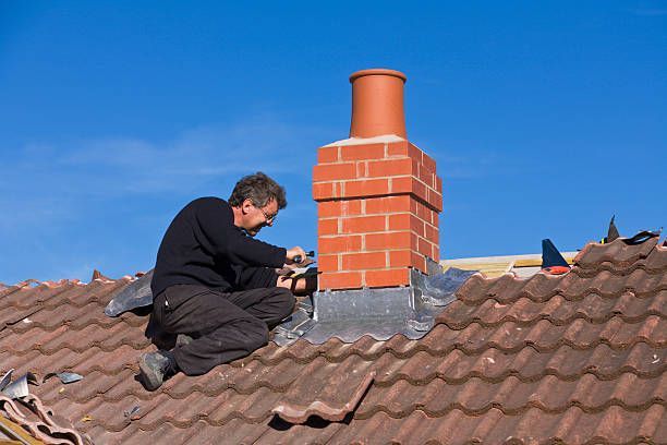 A man is sitting on top of a tiled roof fixing a chimney.