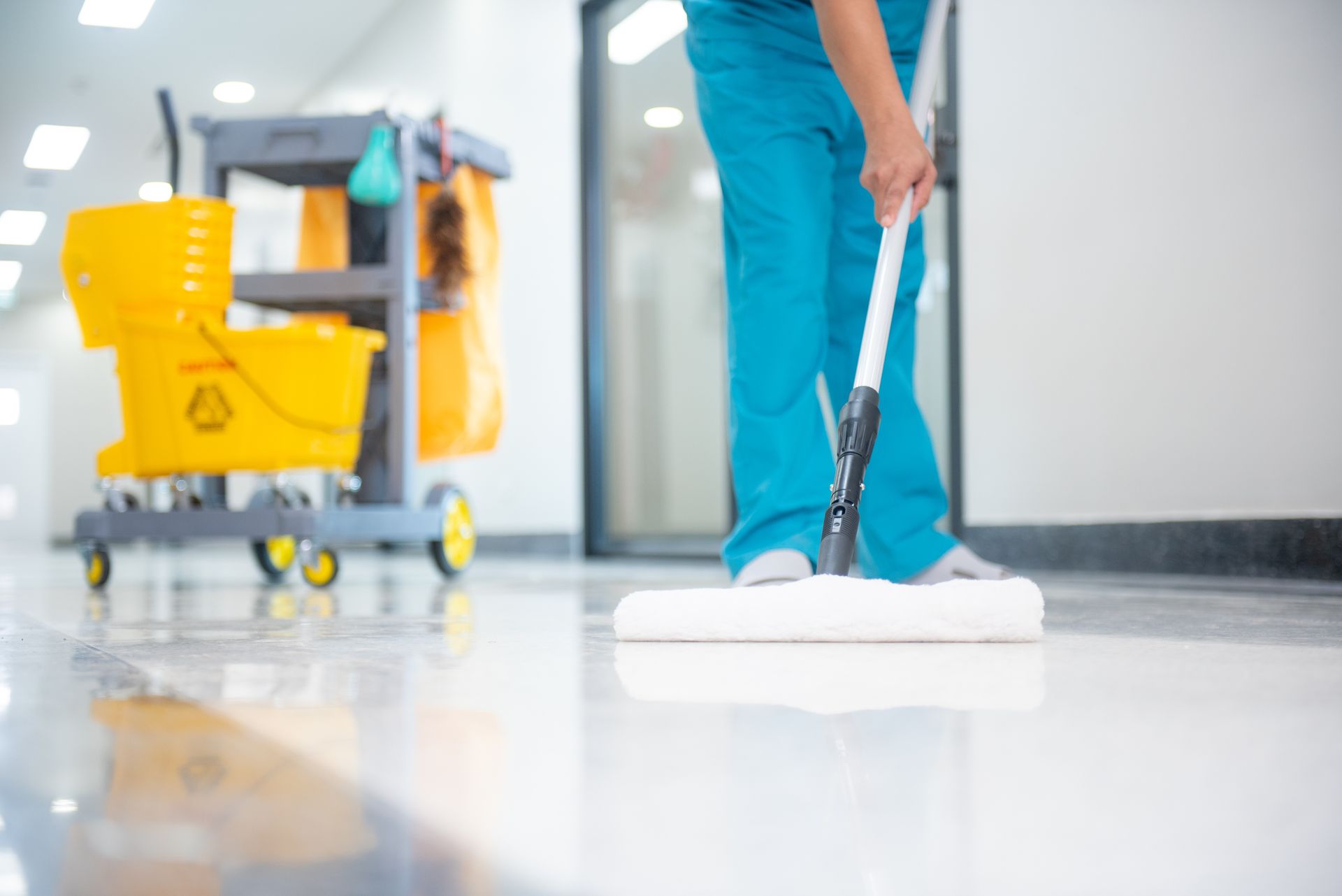 Une personne en blouse bleue lave un sol carrelé de blanc brillant dans un couloir d'hôpital ; un chariot de nettoyage se trouve à proximité.