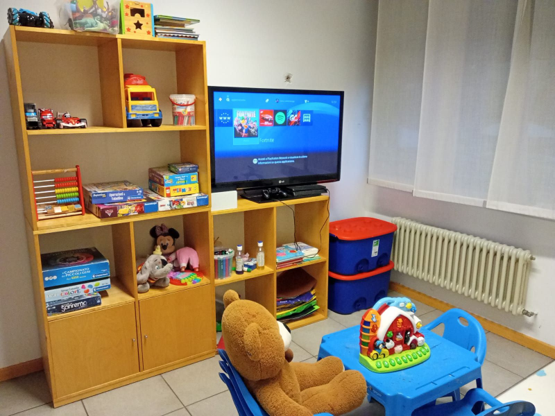 Stanza dei giochi per bambini: scaffali in legno con giocattoli, TV, tavolo/sedie blu, orsacchiotto e un termosifone.