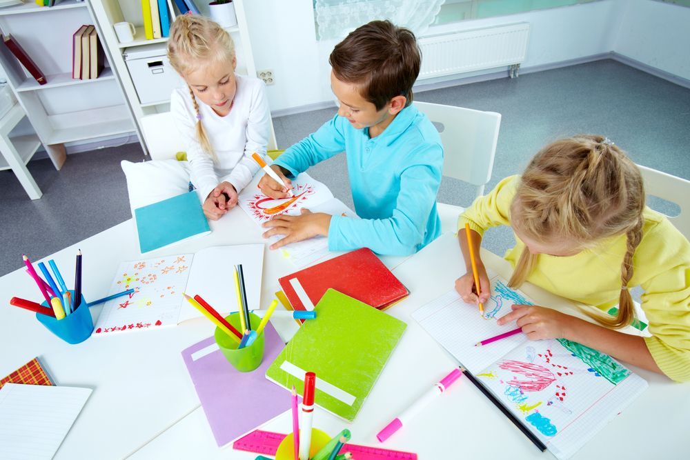 Tre bambini disegnano e colorano seduti a un tavolo bianco in un'aula.