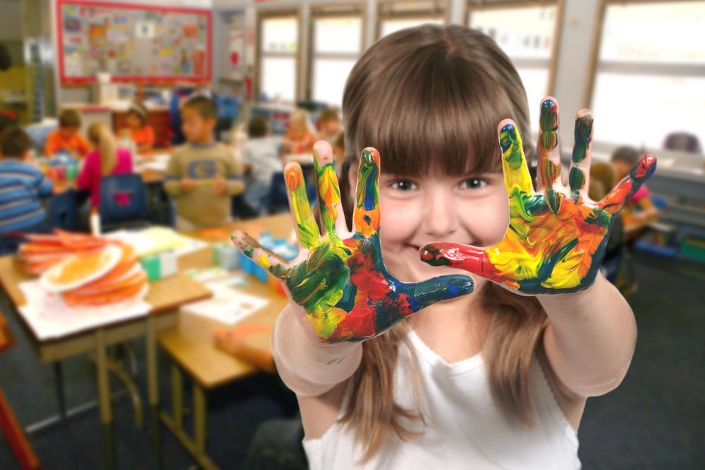 Una bambina sorride, mostrando le mani ricoperte di vernice colorata, in una classe con altri bambini.