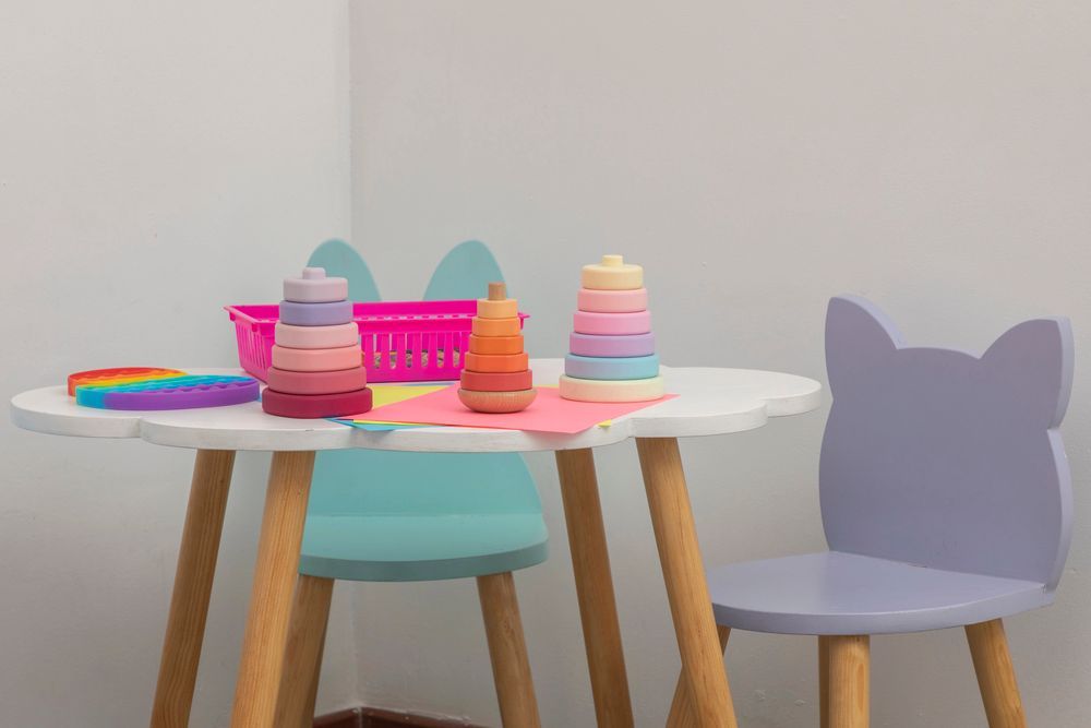 Stanza dei giochi per bambini: scaffali in legno con giocattoli, TV, tavolo/sedie blu, orsacchiotto e un termosifone.