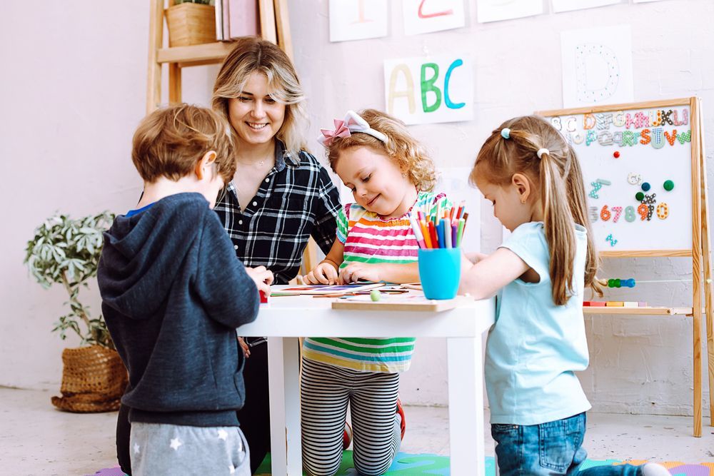 Un'insegnante e tre bambini piccoli seduti a un tavolo bianco colorano con i pastelli in un'aula ben illuminata.