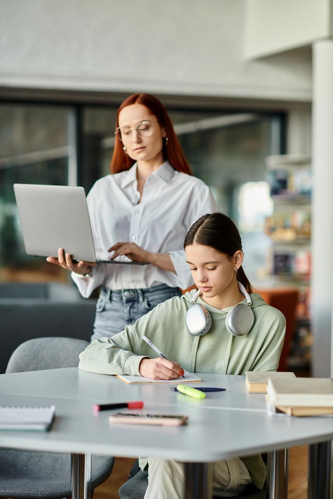 Una donna con i capelli rossi tiene in mano un computer portatile ed è in piedi dietro una persona che scrive a un tavolo, entrambi in una biblioteca.