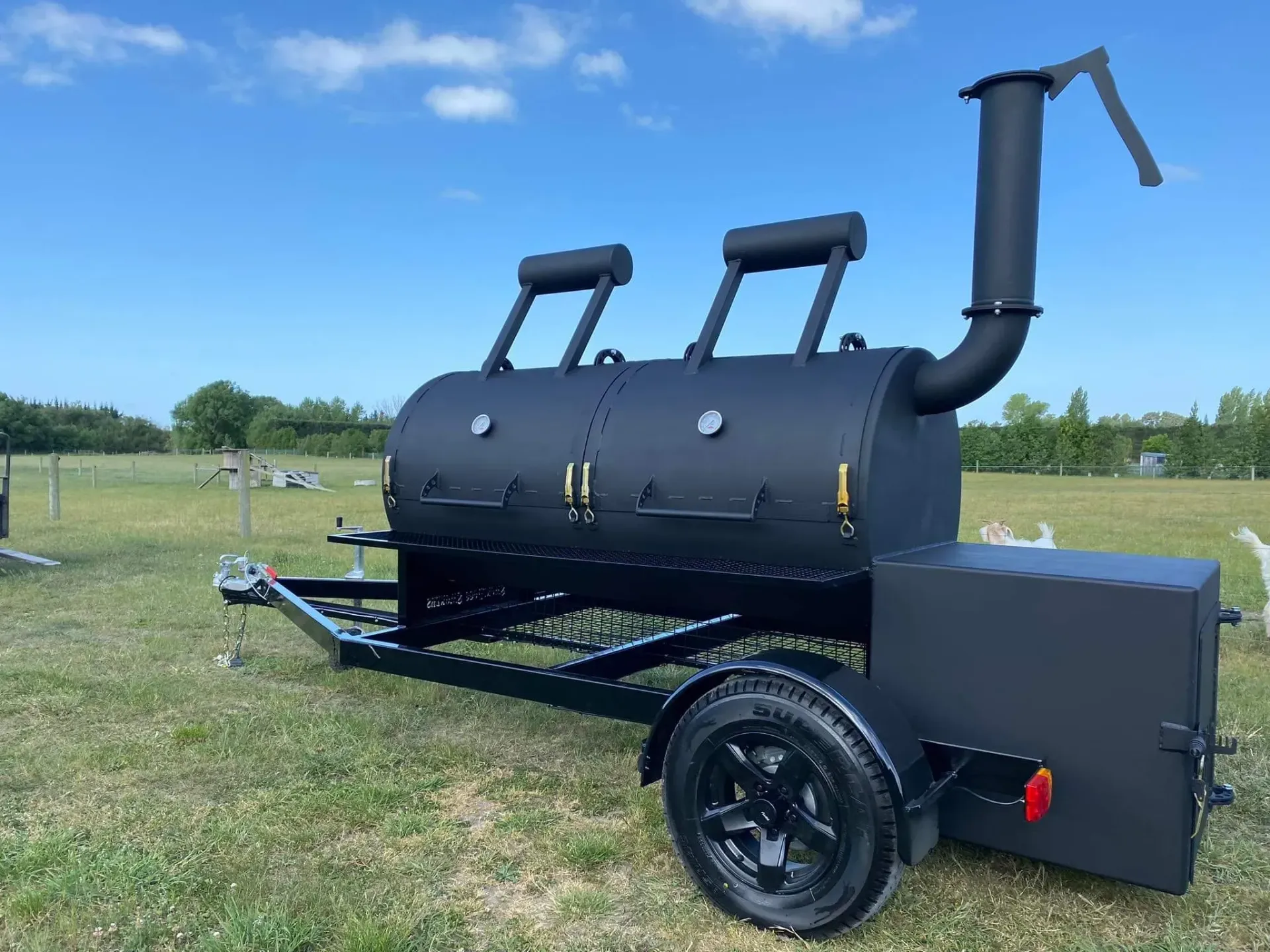 A large black bbq grill is sitting on top of a trailer in a grassy field.