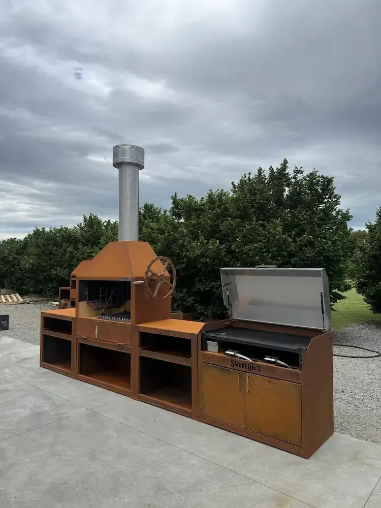 A large, rust-colored outdoor kitchen featuring a wood-burning grill with a chimney and a side gas grill on a patio.