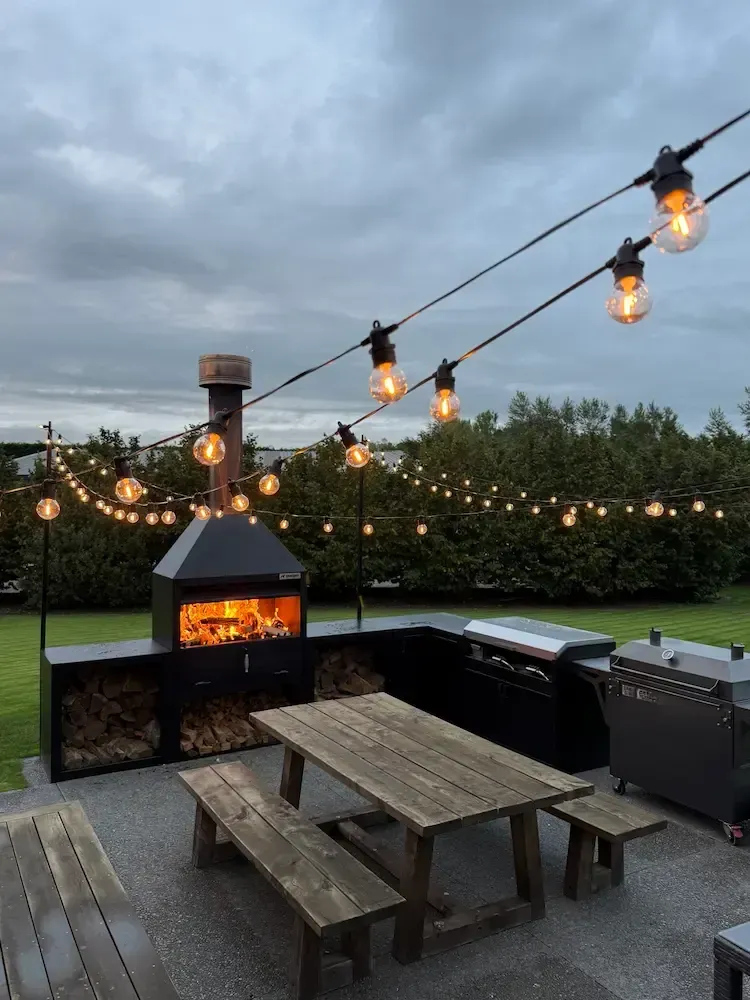 Outdoor patio with a wooden picnic table and benches, a built-in fireplace with fire, and hanging string lights at dusk.