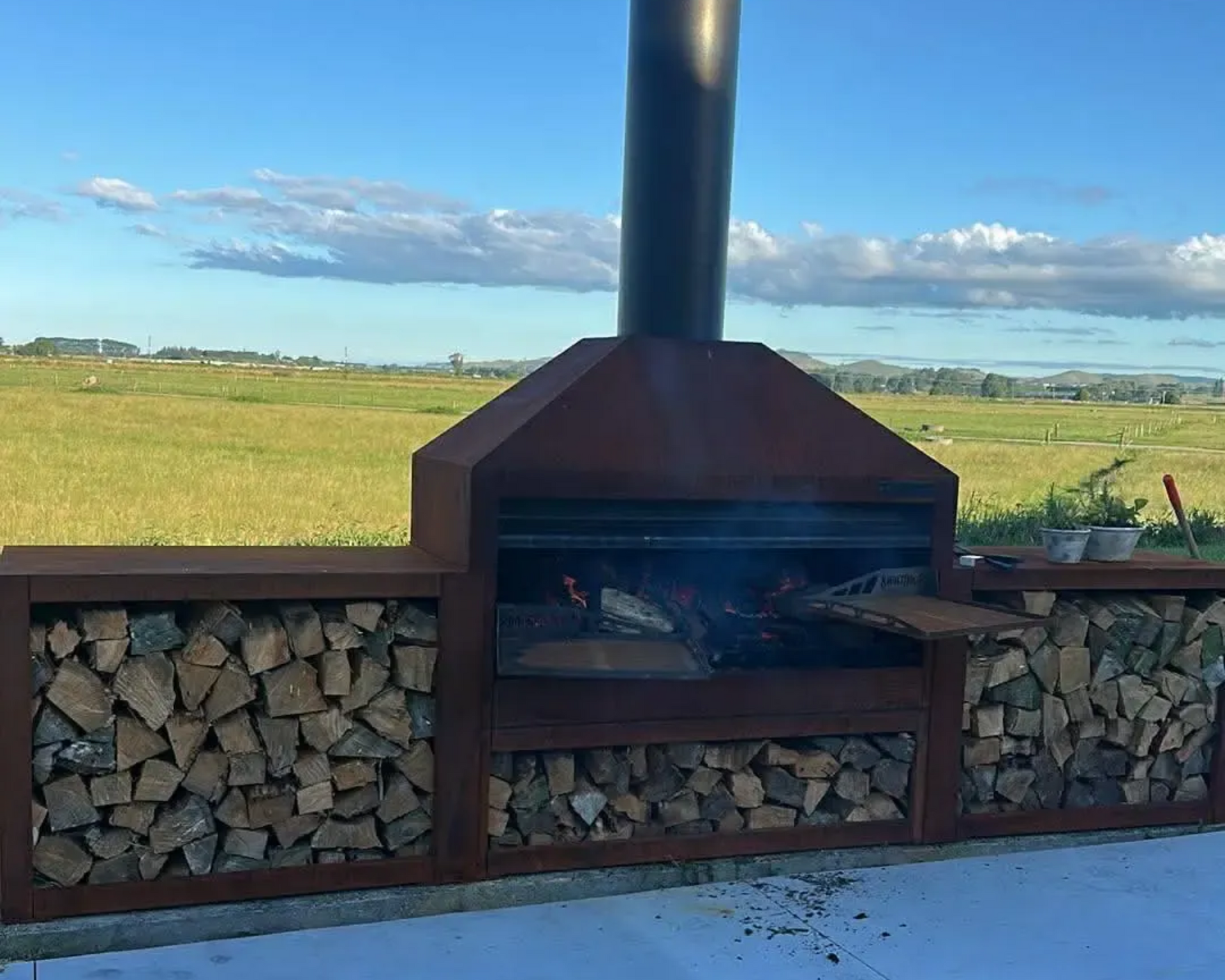 Outdoor grill with wood storage, built into a concrete base. Fire burning, set against a green field and cloudy sky.