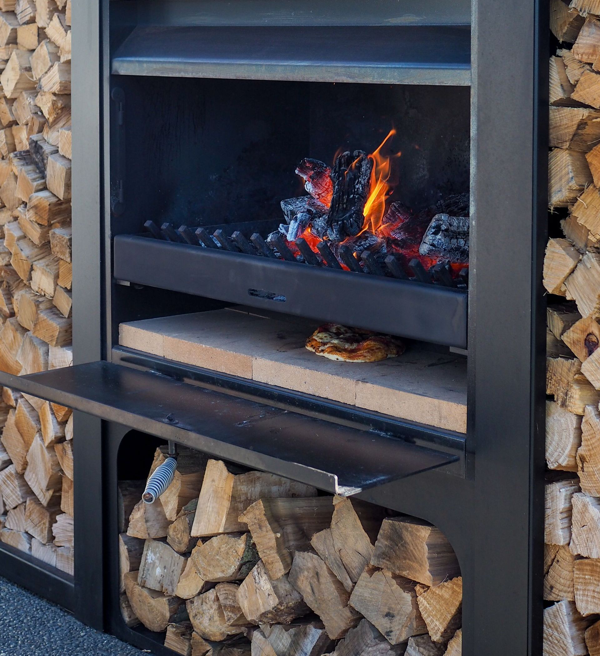 A pizza is cooking in a fireplace next to a pile of wood.