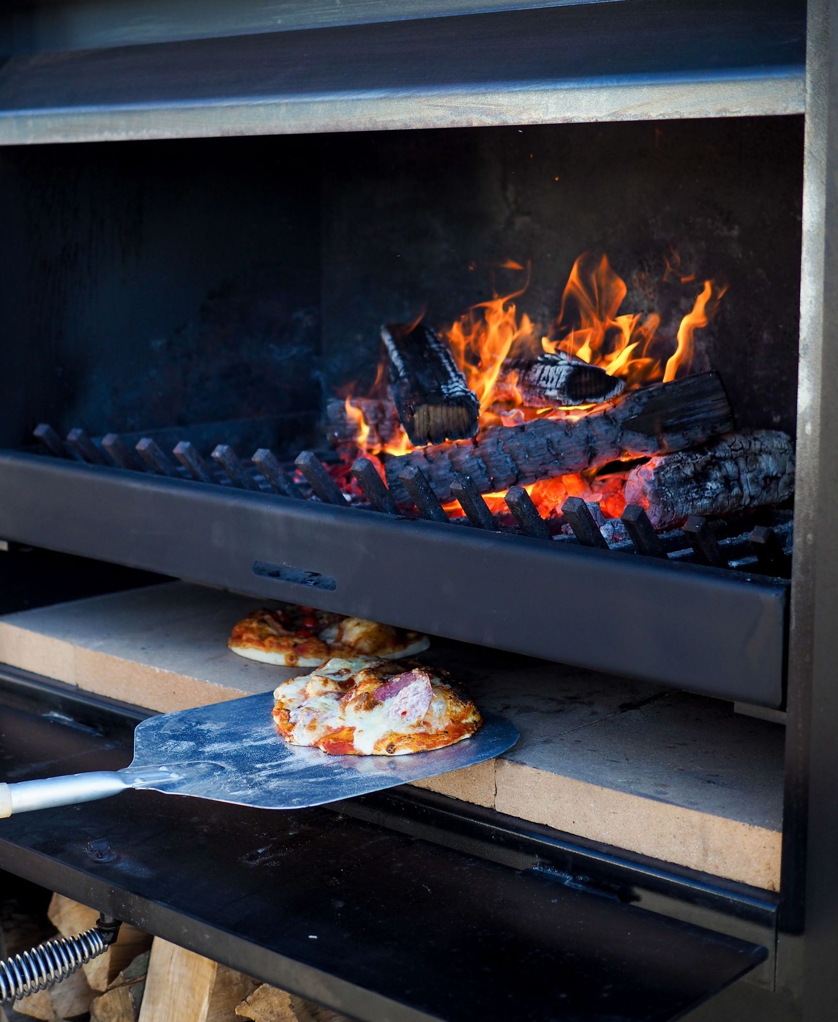 Pizza being removed from a wood-fired oven with flames and a pizza stone.