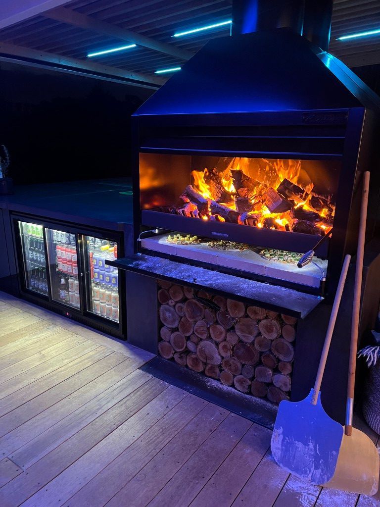 Outdoor wood-fired grill with a fire ablaze, built-in bar fridge, and wood storage. Blue accents illuminate the scene.