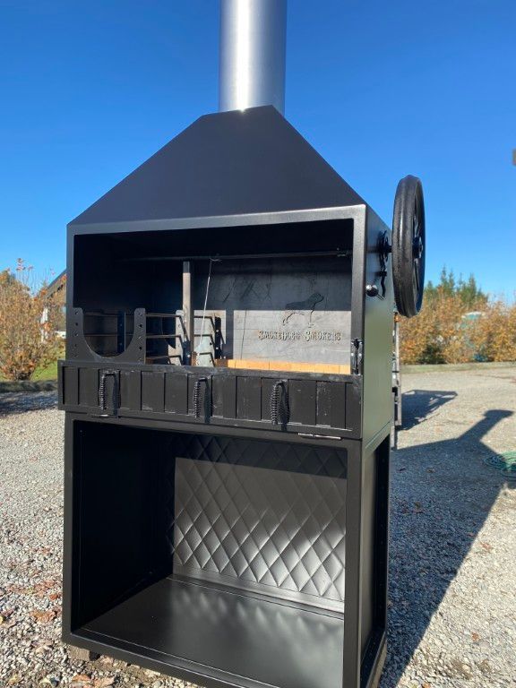 A large grill with a chimney and wheels is sitting on a gravel road.