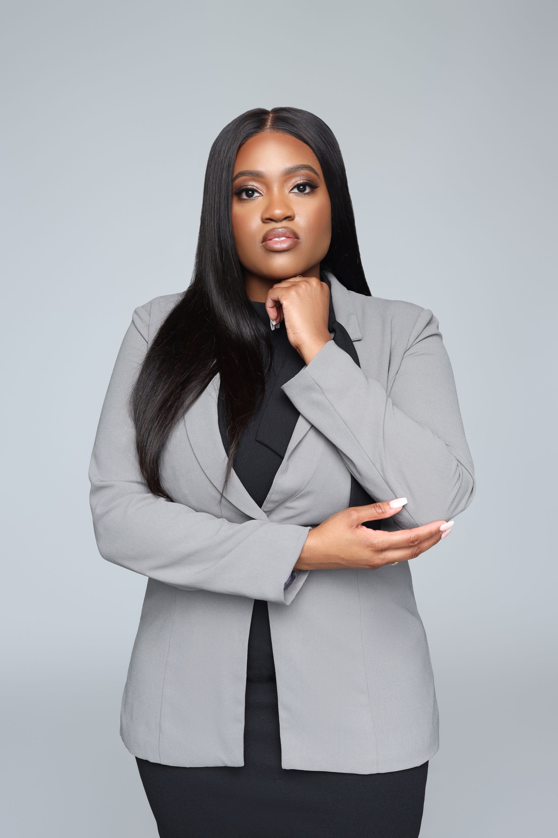 A woman in a gray jacket and black dress is standing with her hands on her chin.