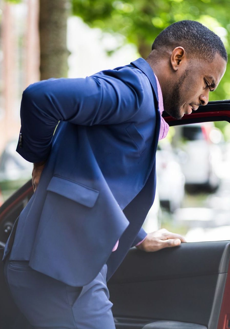 A man in a suit is standing next to a car holding his back.