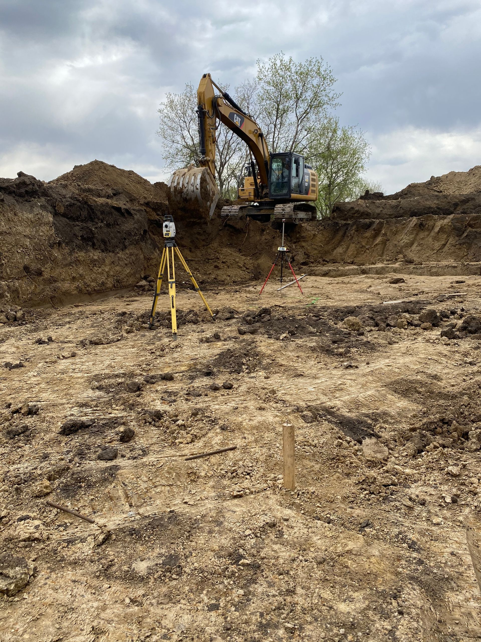 An excavator in a construction site, surrounded by dirt mounds, and surveying equipment. Overcast sky.