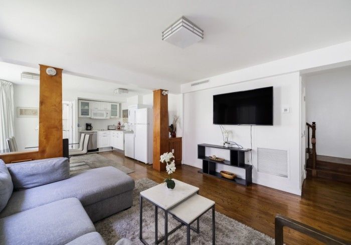 Bright modern living room with gray sofa, wall-mounted TV, kitchen area, and hardwood floors