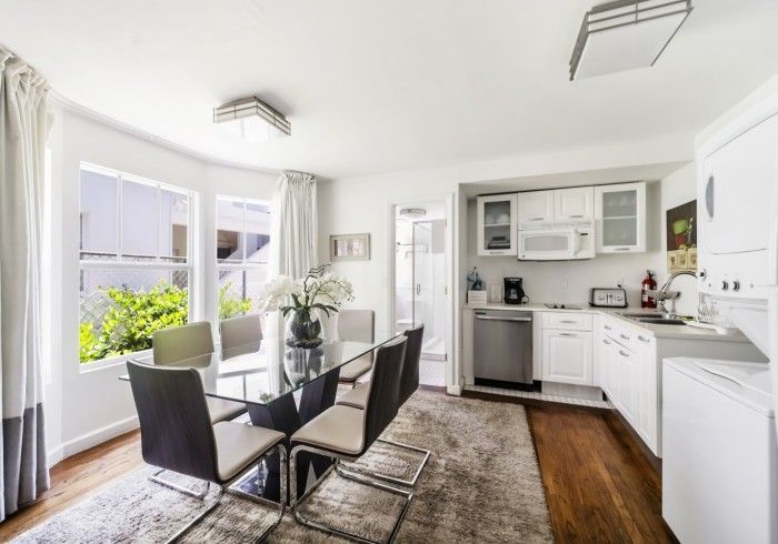 Bright dining area with black chairs, glass table, white kitchen cabinets, and large windows with curtains