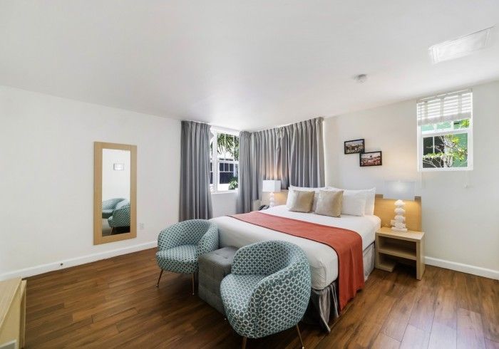 Bright bedroom with a bed, teal chairs, wood floor, gray curtains, and a wall mirror.