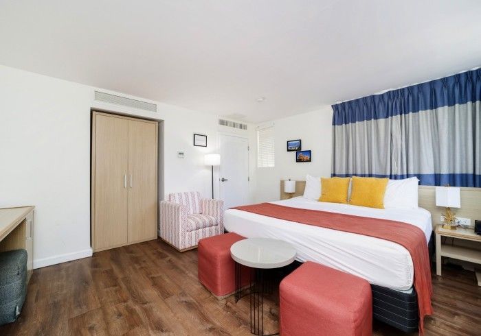 Bright hotel room with a bed, red stools, white table, striped armchair, and blue curtains.