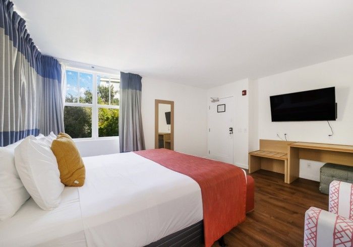 Bright hotel room with a bed, window, desk, wall TV, and blue curtains