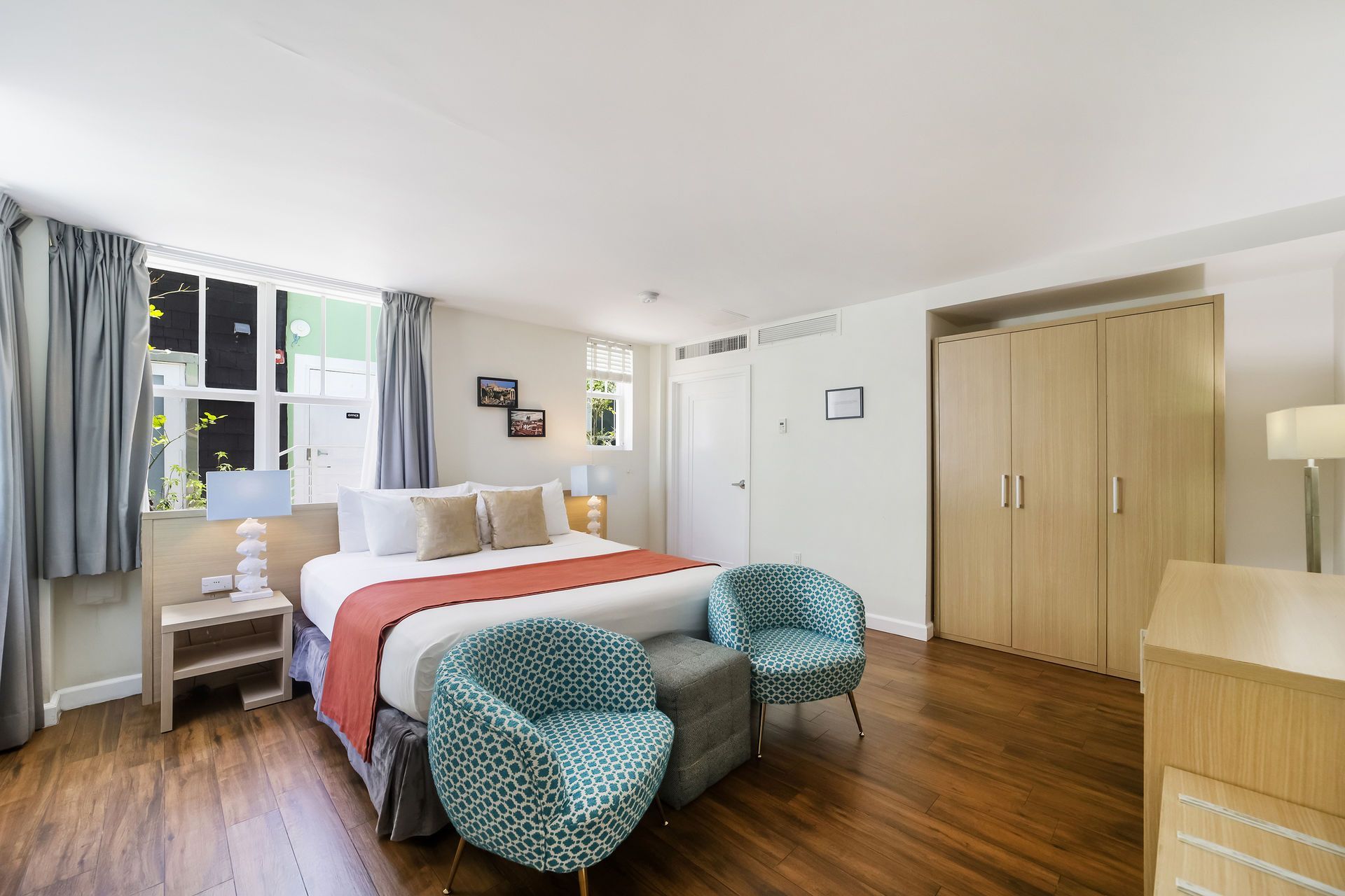 Bright bedroom with a bed, teal chairs, wooden floor, and large windows with curtains.
