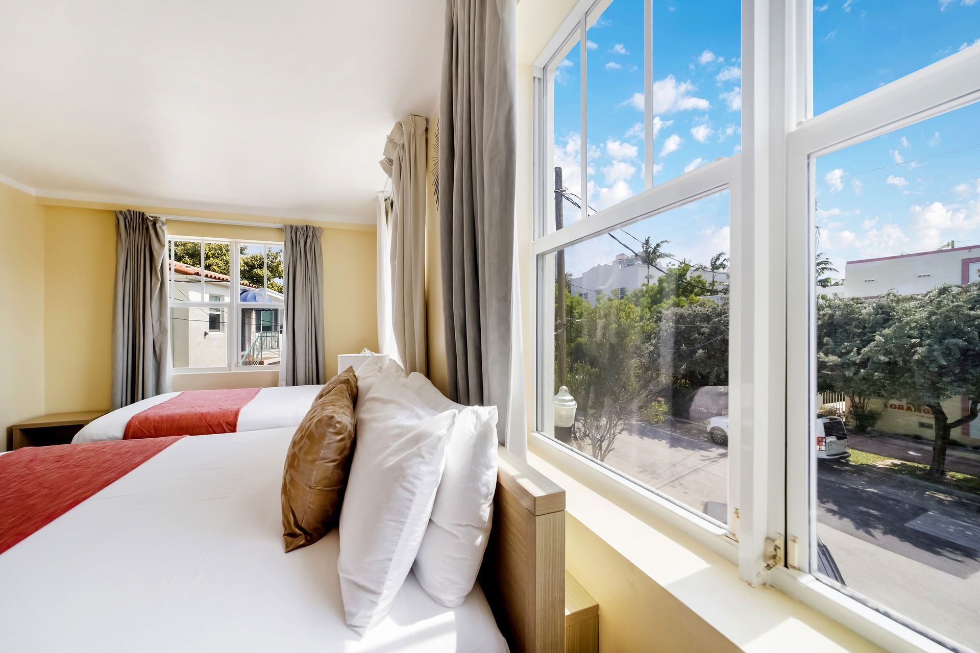 Bright bedroom with twin beds and large windows overlooking a sunny street and trees