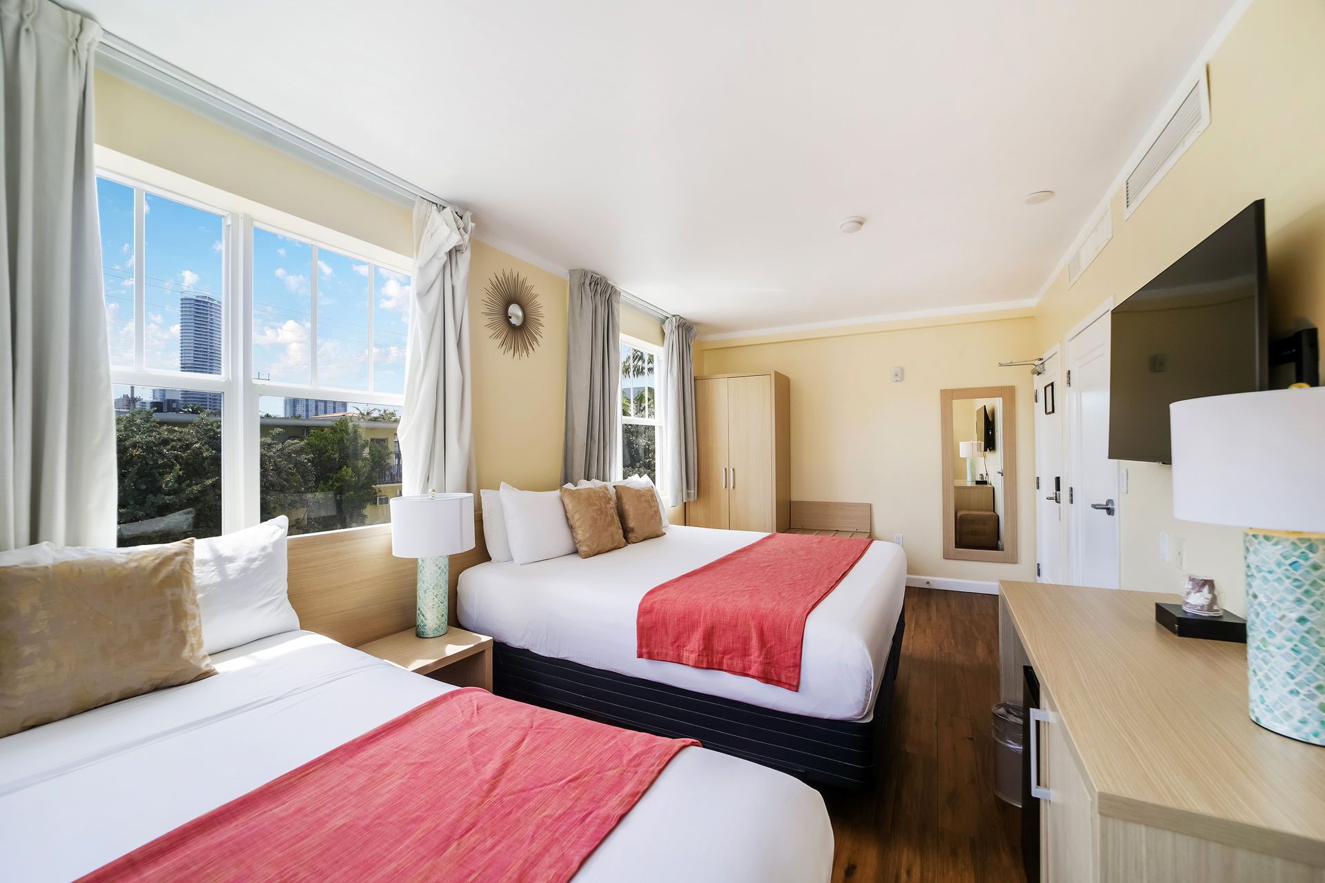 Bright hotel room with two beds, red throws, large windows, and wall-mounted TV