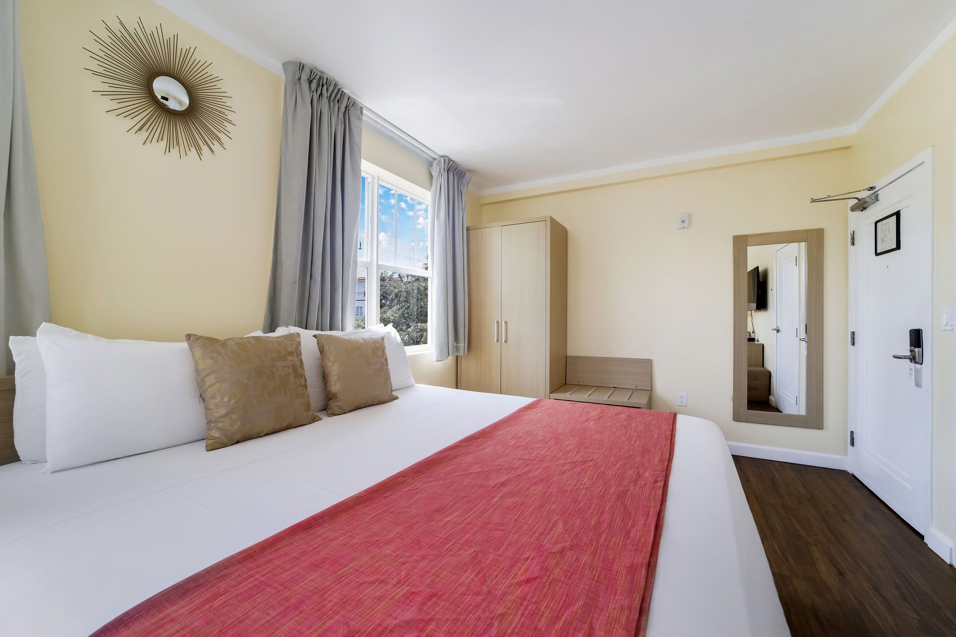 Bright bedroom with white bed, red runner, gray curtains, window, mirror, and wood floor.