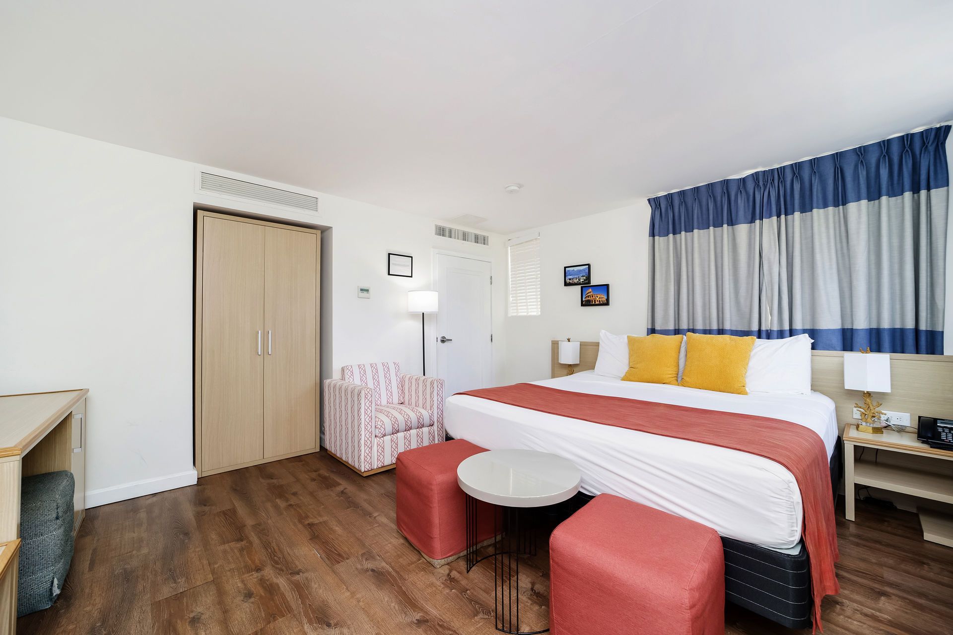 Modern hotel room with a bed, red stools, blue curtains, and wood flooring